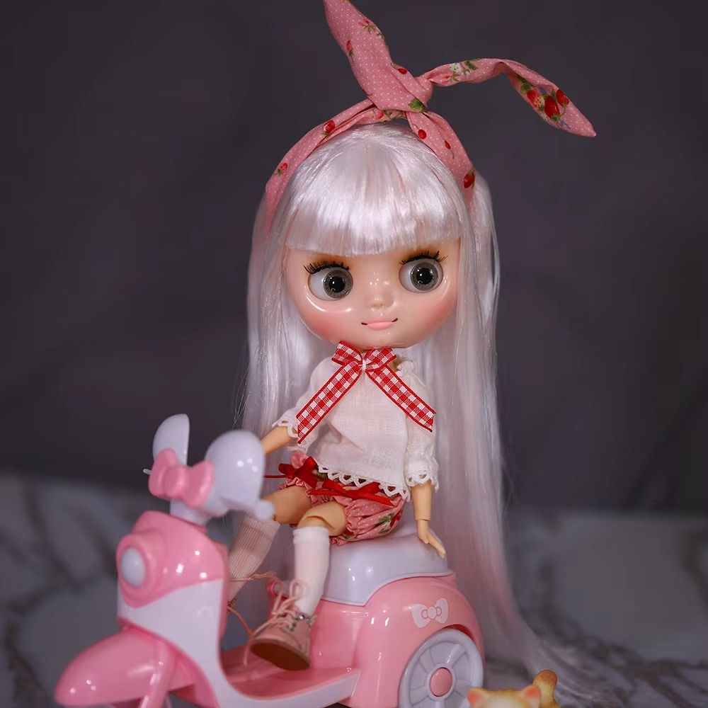 Middie Blythe doll with shiny face and white hair, wearing pink accessories, sitting on a pink scooter.