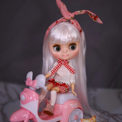 Middie Blythe doll with shiny face and white hair, wearing pink accessories, sitting on a pink scooter.