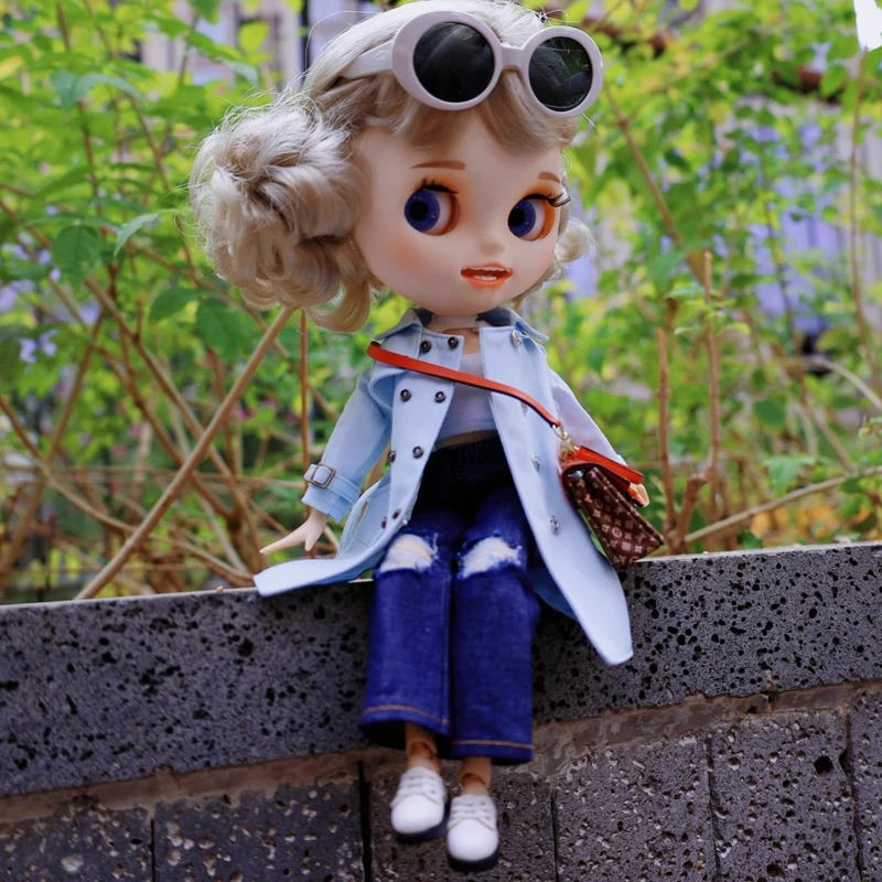 Neo Blythe doll wearing a blue trench coat, stylish jeans, and sunglasses, sitting on a wall surrounded by greenery.