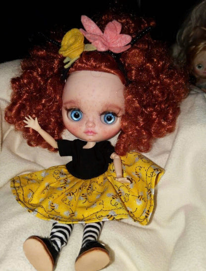 Middie Blythe Doll with red curly hair, blue eyes, and a yellow skirt with black stripes, waving on a soft surface.